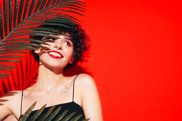 Fashion beauty portrait of young smiling brunette woman with evening stylish  makeup and perfect clean skin. Sexy model with curly short hair posing in studio. With red bright lips. Tropical palm leaf