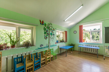 A bright kindergarten room with indoor plants on window sills and colorful furniture.
