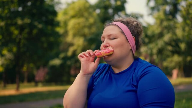 Plus size woman eating donut in park, overeating sweets, extra weight problem