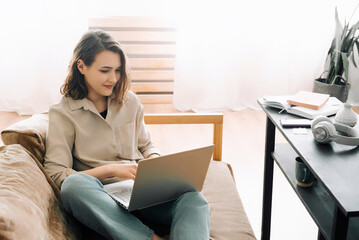 Cheerful millennial woman typing on a laptop, attending a virtual meeting while seated on a sofa with a gadget. Joyful millennial woman typing on a laptop attending a virtual meeting seated on a sofa.