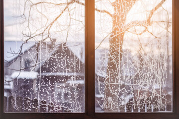 Winter Landscape View from the Window on Snow-covered white Houses and Trees in Winter Countryside. Frosty Pattern on Window  Glass Background.
