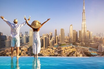 A happy tourist couple on vacation time stands by the pool edge and enjoys the panoramic sunset view of the Dubai city skyline, UAE