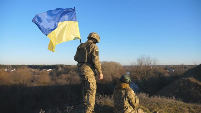 Military couple of ukrainian army sitting on lawn with Ukraine flag at countryside. Young soldiers in camouflage uniform with national banner resting outdoor. Resistance to russian invasion. Dollyshot