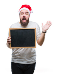 Young caucasian hipster man wearing christmas hat holding blackboard over isolated background very happy and excited, winner expression celebrating victory screaming with big smile and raised hands