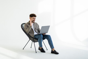 Portrait of attractive man sitting in chair using laptop calling contact over white background