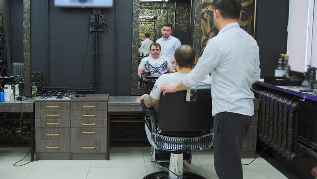 A man explains to a hairdresser how he wants to get a haircut while sitting on an armchair in a beauty salon. Medium Long Shot