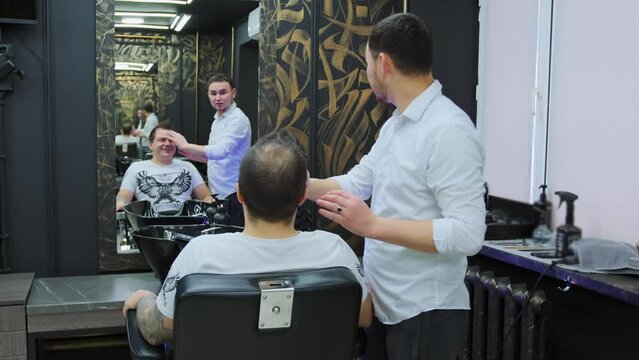 The hairdresser discusses with the client how he wants to get a haircut while sitting in an armchair in a beauty salon. The stylist looks at what type of hair a man has before cutting