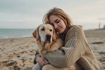A young woman in a sweater hugs a golden retriever dog sitting on the sand near the sea