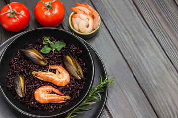 Shrimps and mussels with black rice on plate. Tomatoes, shrimps in bowl. Dark wooden background