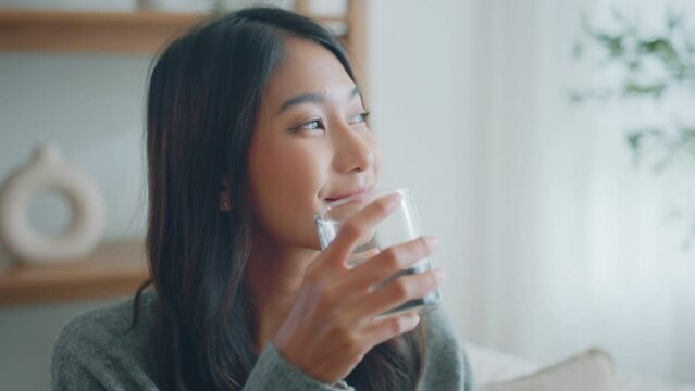 Asian woman drink water start new day with healthy life habit, Female holding glass drinking clean mineral natural still water in living room at home morning, Lifestyle healthcare concept