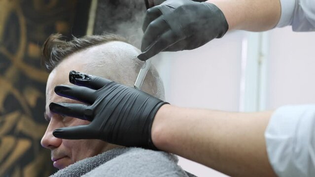 A man in a barbershop gets a fashionable hairstyle on his head, his skin is shaved bald with a straight razor under a stream of hot steam from a vaporizer. Close-up.