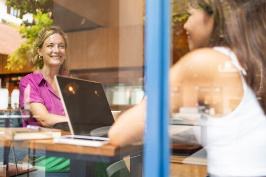 Through the window woman in a vibrant purple shirt enjoys remote working on her laptop from a cafe with her multiethnic colleague in Chiang Mai, Thailand - Powered by Adobe