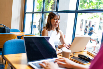 A cheerful Asian woman working on a laptop in a bright cafe, with a Caucasian woman during remote work environment in Chiang Mai, Thailand