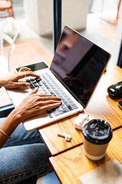Cropped unrecognizable woman's hands typing on a laptop in a cafe, with a smartphone, earphones, and a coffee cup on the table during remote work in Chiang Mai, Thailand