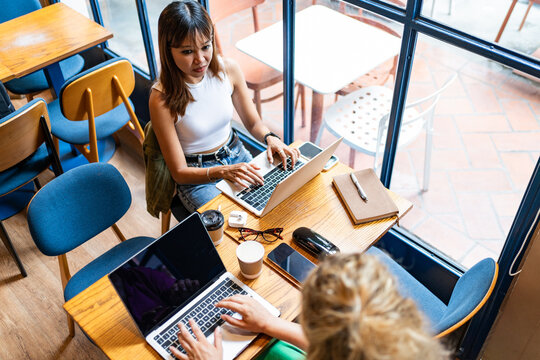 From Above Asian Woman In A White Top Working On Her Laptop In A Cafe With A City View, Engaging In Remote Work In Chiang Mai, Thailand, With A Partially Visible Colleague Beside Herdigitak