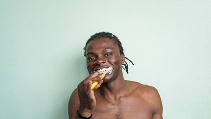 A shirtless black man with a nose ring and earrings brushes his teeth against a light green background