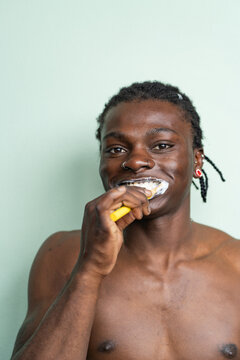 Portrait shirtless black man with a nose ring and earrings brushes his teeth against a light green background