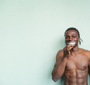 A shirtless black man with a nose ring and earrings brushes his teeth against a light green background