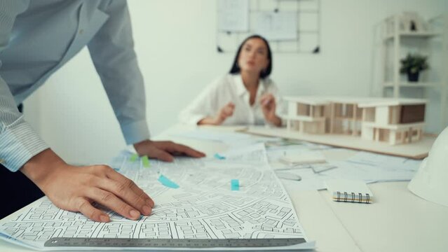 Skilled caucasian interior designer working with house model while discussing with asian architect engineer about house plan on meeting table with blueprint and house model scatter around. Immaculate.