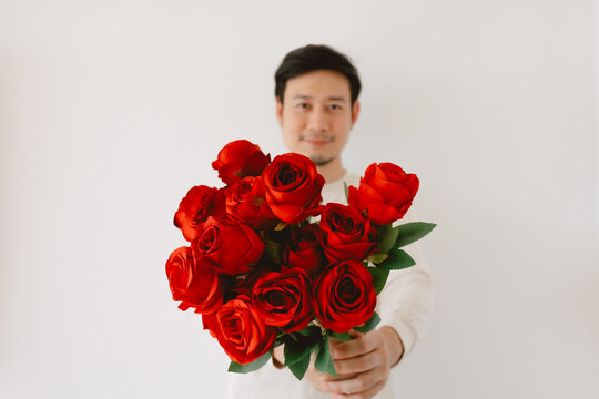Handsome Asian Man holding red roses and giving flowers on Valentines day, happy smiling and looking to camera, standing isolated over white background wall. 