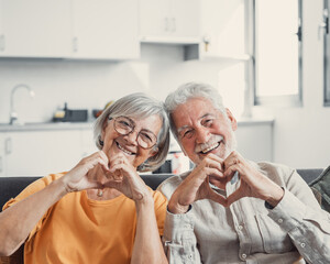 Close up portrait happy sincere middle aged elderly retired family couple making heart gesture with fingers, showing love or demonstrating sincere feelings together indoors, looking at camera..