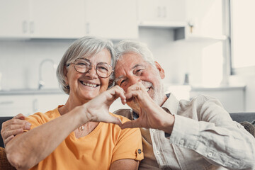 Close up portrait happy sincere middle aged elderly retired family couple making heart gesture with fingers, showing love or demonstrating sincere feelings together indoors, looking at camera.. - Powered by Adobe