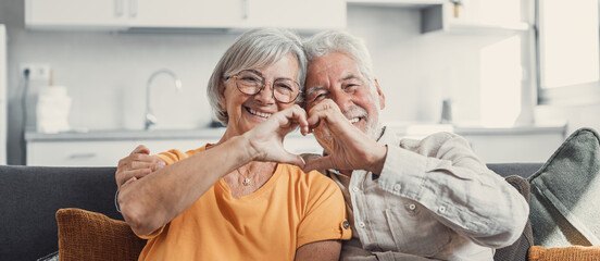 Close up portrait happy sincere middle aged elderly retired family couple making heart gesture with fingers, showing love or demonstrating sincere feelings together indoors, looking at camera.. - Powered by Adobe