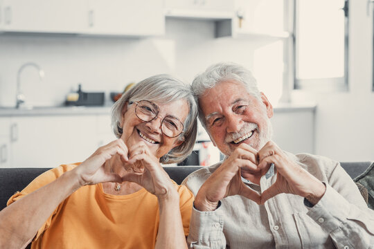 Close Up Portrait Happy Sincere Middle Aged Elderly Retired Family Couple Making Heart Gesture With Fingers, Showing Love Or Demonstrating Sincere Feelings Together Indoors, Looking At Camera..