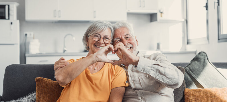 Close Up Portrait Happy Sincere Middle Aged Elderly Retired Family Couple Making Heart Gesture With Fingers, Showing Love Or Demonstrating Sincere Feelings Together Indoors, Looking At Camera..