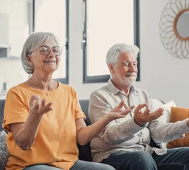 Poster Calm senior middle aged couple practicing yoga together sitting in lotus pose on sofa, mindful peaceful mature man and woman meditating relaxing in living room at home, old people healthy lifestyle. © Daniel