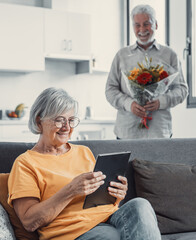 Obraz premium Old man giving flowers at his wife sitting on the sofa at home for the San Valentines’ day. Pensioners enjoying surprise together. In love people having fun..