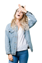 Beautiful young blonde woman wearing denim jacket over isolated background surprised with hand on...