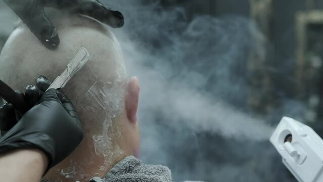 The barber's hands in black gloves shave the visitor's head bald, making him a stylish hairstyle with a straight razor under a stream of hot steam from a vaporizer.