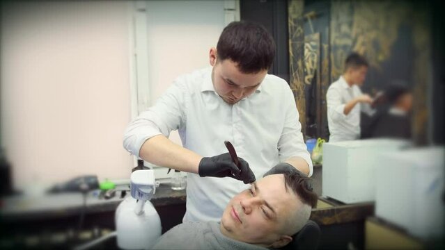 An Asian hairdresser shaves his head with a straight razor under a stream of hot steam
