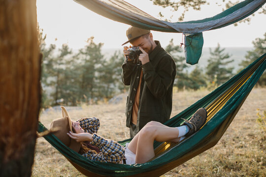 Couple having fun relaxing in hammocks outdoors. A guy takes a picture of a girl on a film camera.