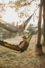 A guy and a girl are relaxing in hammocks in a pine forest at sunset.