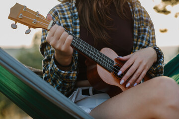 A young girl plays the ukulele while sitting in a hammock at sunset. Close-up.