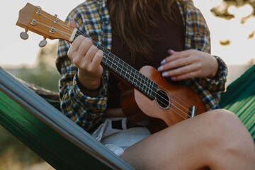 A young girl plays the ukulele while sitting in a hammock at sunset. Close-up.
