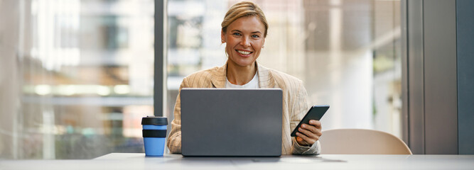 Smiling female entrepreneur use phone while working on laptop in modern office background