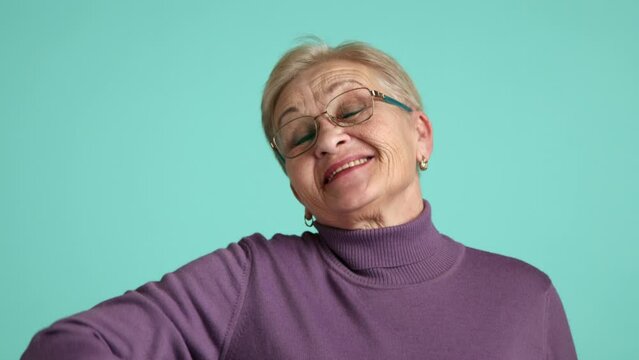 Cheerful senior lady in spectacles with blonde hair, short haircut, wearing purple turtleneck standing over blue background, waving her hand, smiling. High quality 4k footage