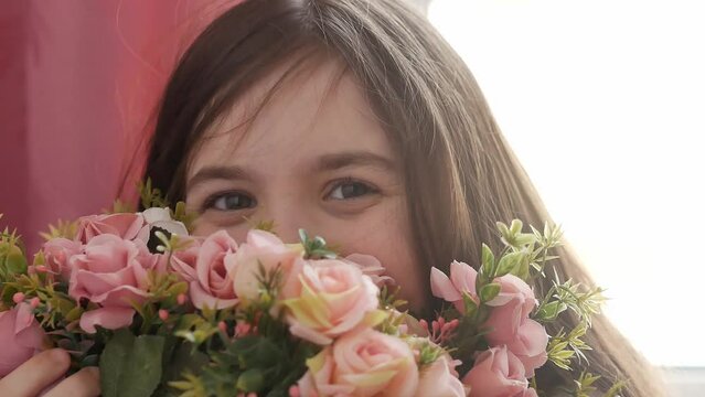 A little girl is smiling, looking into the frame, peeking out from behind a bouquet of flowers made of small white and delicate pink roses. Happiness, the joy of a gift.