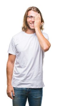 Young handsome man with long hair wearing glasses over isolated background covering one eye with hand with confident smile on face and surprise emotion.
