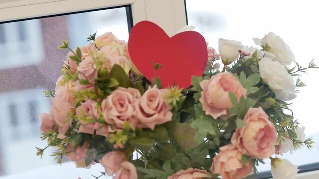 A beautiful bouquet of white and pink wild flowers in small buds and a postcard in the shape of a red heart made of paper. A gift, decoration of the wedding table of the hall in celebration.