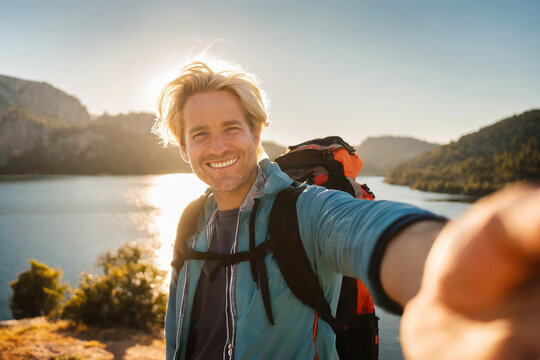 Ein m&auml;nnlicher, blonder Backpacker macht ein Selfie und l&auml;chelt in die Kamera, Sonnenuntergang und See im Hintergrund
