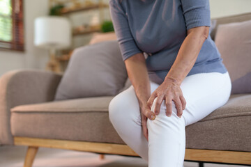 Old woman with knee pain sits on the sofa. Senile woman with body aches, leg pain sitting on a sofa in the living room stretching. - obrazy, fototapety, plakaty