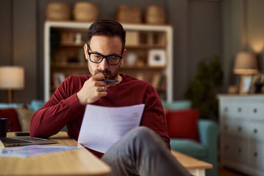 A focused male editor revising a draft work on a paper that he is holding while sitting in his home office.
