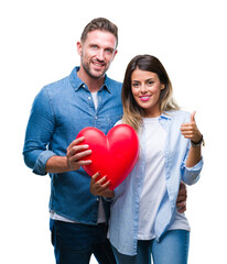 Young couple in love holding red heart over isolated background happy with big smile doing ok sign, thumb up with fingers, excellent sign