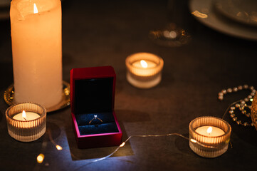 Engagement ring in a red box. Burning candles for a surprise marriage proposal. Decoration candle. Details closeup. Luxury candlelight date in restaurant. Table setting for couple on Valentine's day.