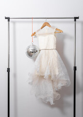 White puffy dress hangs on hanger on clothes rail with disco ball on white wall background.