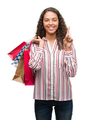 Young hispanic woman holding shopping bags surprised with an idea or question pointing finger with happy face, number one